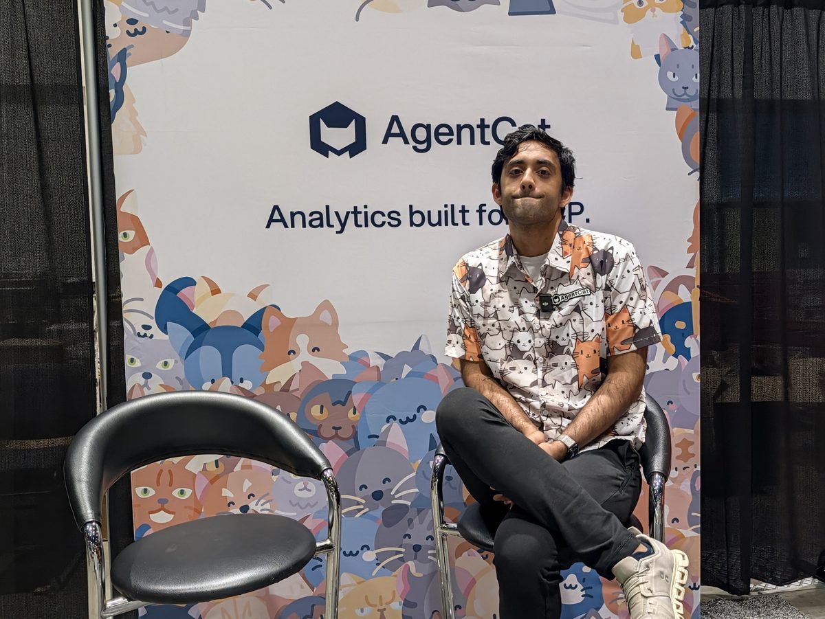 Kashish Hora sitting on a stool in front of the AgentCat booth wall, Analytics built for MCP, decorated with cartoon cats