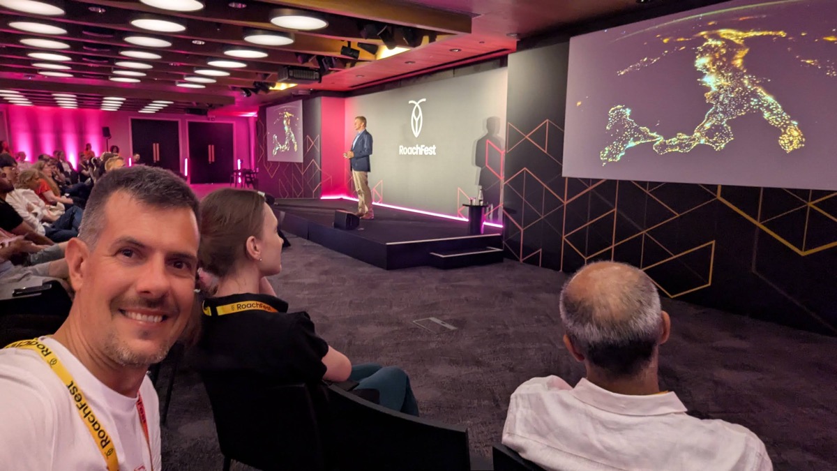 The RoachFest London main stage, a speaker in front of a screen showing city lights from orbit, with the audience seated in a pink-lit room